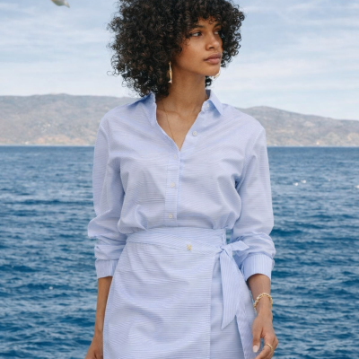Mulher a usar vestido riscado azul e branco com cinto laço, em frente ao mar e céu azul com gaivota.