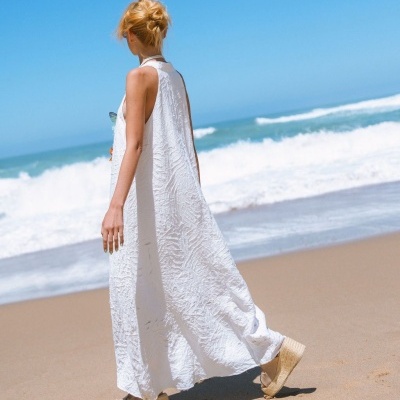 Mulher de vestido branco longo e sandálias de cunha na praia com mar e céu azul.