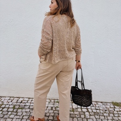 Mulher de costas com casaco de malha bege, calças largas bege, sandálias castanhas, segurando bolsa preta sobre calçada e parede branca.