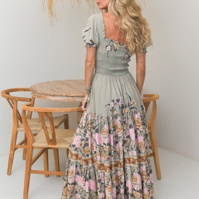 Mulher de costas a usar vestido longo floral verde com rosa e castanho, em sala com mesa e cadeiras de madeira e ramos decorativos secos