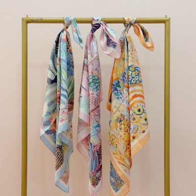 Três lenços florais coloridos pendurados numa barra metálica amarela com fundo rosa claro.