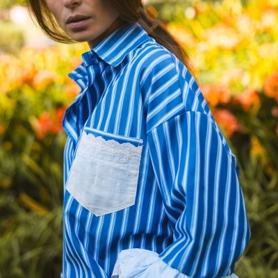 Camisa feminina azul e branca com bolso branco e renda, com flores ao fundo.