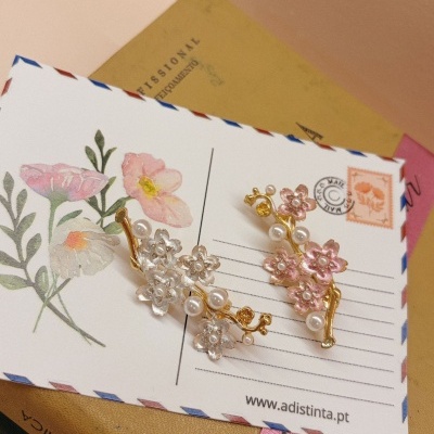 Brincos florais dourados com flores brancas e rosa e pérolas sobre cartão decorativo com flores.