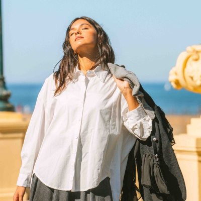 Mulher com camisa branca, saia cinza e casaco preto ao ar livre com céu e mar ao fundo.