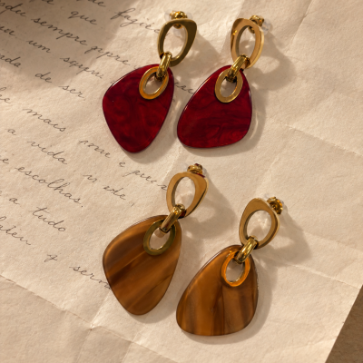 Dois pares de brincos com pendentes vermelho e castanho sobre papel envelhecido com texto manuscrito.