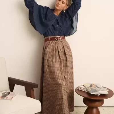 Mulher com camisa azul, saia xadrez e sapatos castanhos junto a cadeira e mesa com revistas