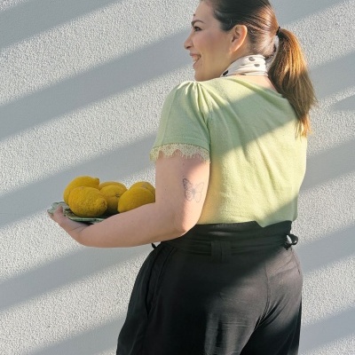 Mulher sorridente de perfil com rabo de cavalo, vestindo camisola verde clara e calças pretas, segurando prato com limões amarelos.