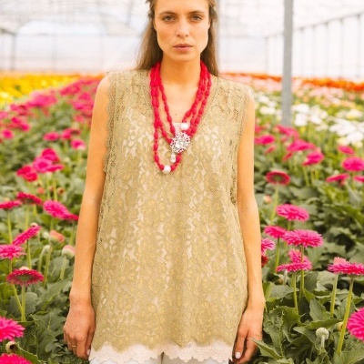 Mulher com blusa de renda bege e colar vermelho em estufa com flores cor-de-rosa.