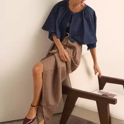Mulher sentada com capa de ganga azul, saia xadrez e sapatos cor vinho.