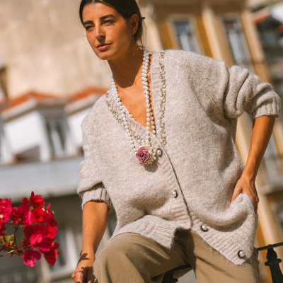Mulher com cardigan bege e colar de pérolas sentada num corrimão com flores vermelhas ao lado