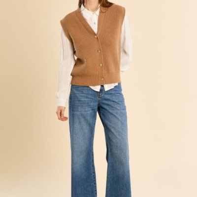 Mulher com camisa branca, colete castanho de malha, jeans azuis largos e botas castanhas de salto.