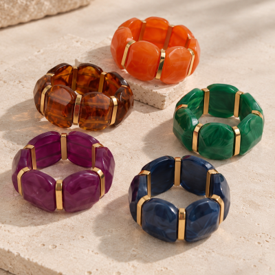 Cinco pulseiras largas coloridas com detalhes dourados sobre pedra clara.