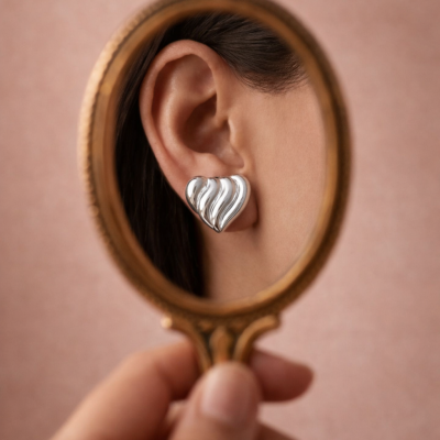 Brincos prateados em forma de coração refletidos num espelho com moldura dourada.