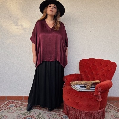 Mulher com blusa vinho acetinada e saia preta ao lado de cadeira vermelha estofada com livros.