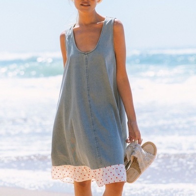 Mulher a usar vestido azul claro com bainha branca às bolinhas vermelhas e óculos de sol vermelhos na praia, segurando sandálias com pérolas.
