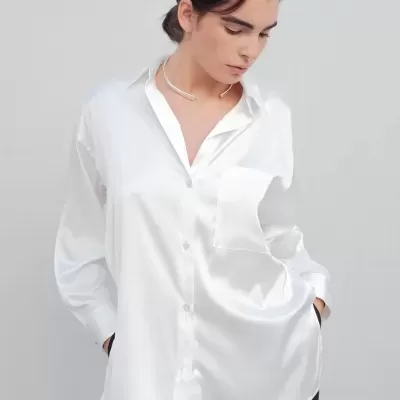 Camisa branca acetinada com bolso e padrão listrado subtil, calças pretas, colar prateado e cabelo apanhado.