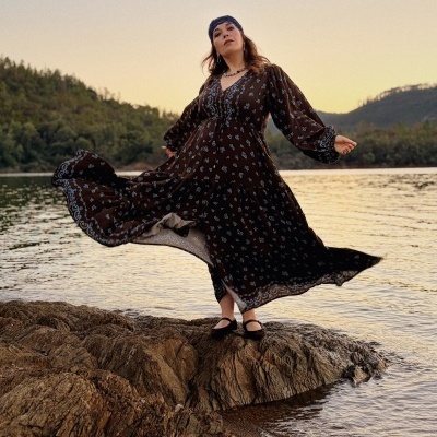 Mulher com vestido comprido castanho de flores azuis junto a lago e colinas ao pôr do sol