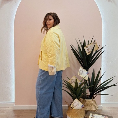 Mulher com casaco amarelo e calças de ganga, em fundo decorativo com plantas e cestos