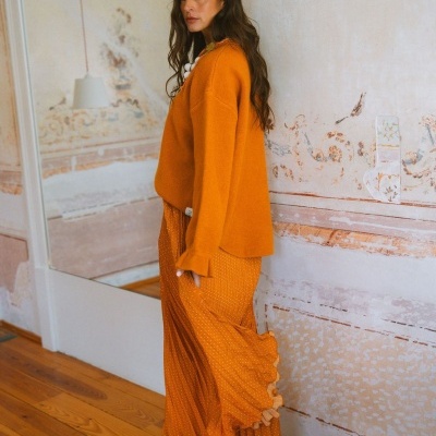 Mulher de cabelo castanho comprido com camisola e saia castanhas alaranjadas em ambiente interior