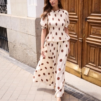 Mulher a usar conjunto creme com bolas castanhas, blusa de manga curta e calças largas, em ambiente urbano com porta de madeira.