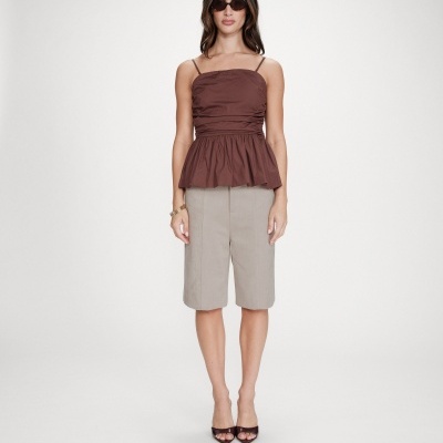 Mulher com blusa castanha de alças, bermuda cinzenta, sapatos pretos e óculos escuros em fundo branco