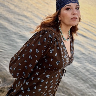 Mulher com vestido de flores castanho e lenço azul ao pôr do sol junto à praia
