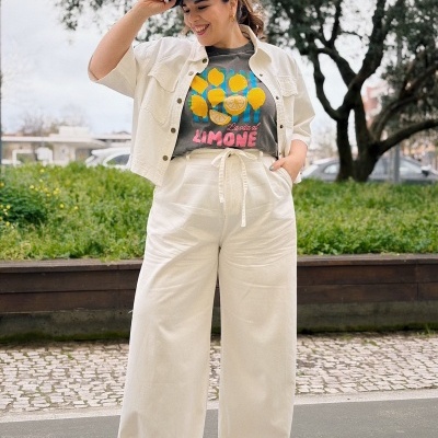 Mulher a usar casaco creme, t-shirt cinzenta com limões e texto, calças largas creme e boné azul ao ar livre