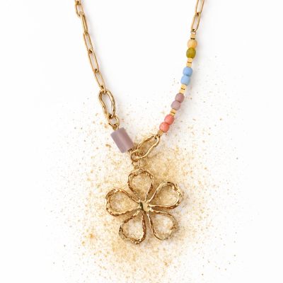 Colar dourado com pendente de flor e contas coloridas sobre fundo branco com pó dourado.