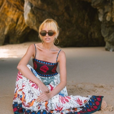 Mulher de vestido estampado sentada na areia em cenário de praia com rochas