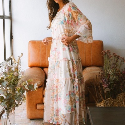Vestido comprido branco com padrão floral multicolorido e cintinho, em ambiente interior com sofá castanho e flores secas