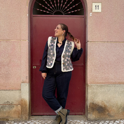 Mulher com colete floral e calças pretas em frente a porta vermelha com número 11 na parede