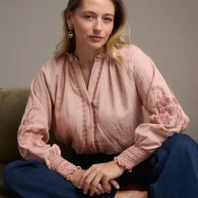 Blusa rosa com bordados florais nas mangas e calças de ganga azuis sentada num sofá verde