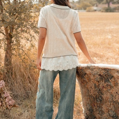 Pessoa de costas com blusa bege e renda branca, calças jeans largas e sapatos, junto a cadeira floral num campo seco.