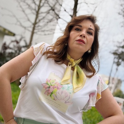 Mulher de t-shirt branca com flor e texto, folhos nas mangas, calças verde-água e lenço amarelo ao pescoço