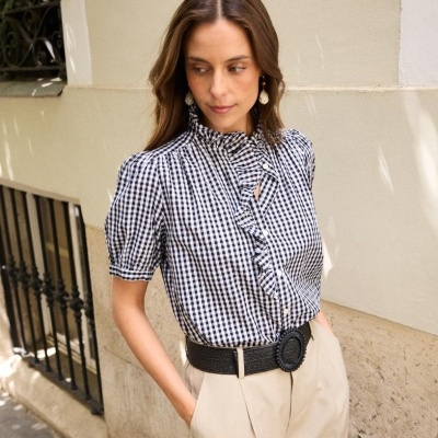 Mulher com blusa xadrez preta e branca de folhos e calças bege com cinto preto junto a parede clara