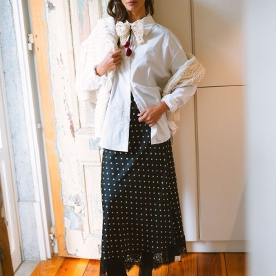 Mulher com camisa branca, saia preta de bolinhas brancas e botas pretas em ambiente interior