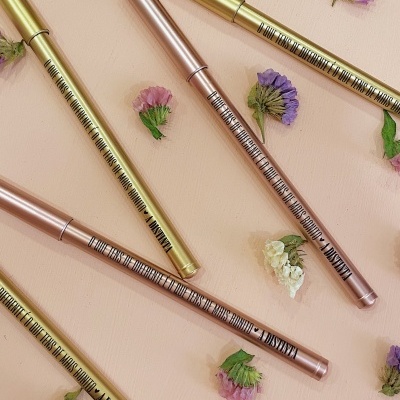 Lápis delineadores dourados e rosa com texto preto sobre superfície rosa clara com flores pequenas coloridas.