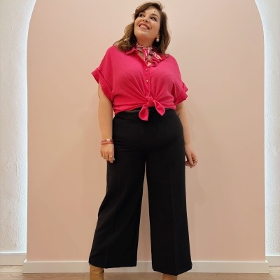 Mulher com camisa fúcsia e calças pretas num interior com parede rosa clara e chão de madeira.