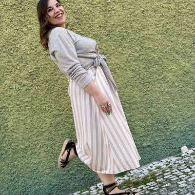 Mulher com blusa cinza, saia riscada bege e branca e sandálias pretas numa calçada de pedras junto a parede verde