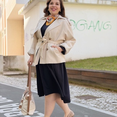 Mulher de casaco bege, vestido preto e sapatos beges numa rua urbana com parede branca e graffiti verde.