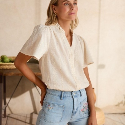 Mulher com blusa branca bordada e jeans azul claro com bordados florais nos bolsos frente, em ambiente interior neutro.