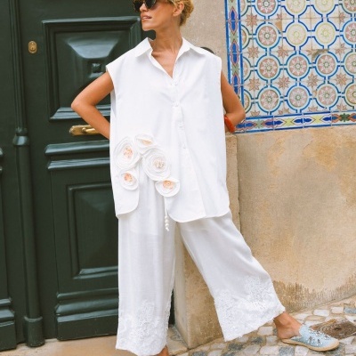 Conjunto feminino branco com camisa sem mangas e calças largas com renda, sapatilhas azuis, posando junto a porta verde e azulejos coloridos.
