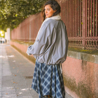 Mulher com casaco cinza claro, saia xadrez azul, botas bege e cachecol branco num passeio urbano