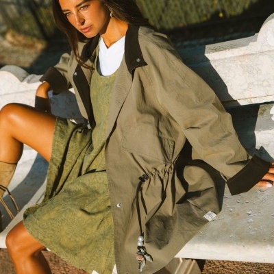 Mulher sentada num banco de pedra com vestido verde e casaco bege e castanho.