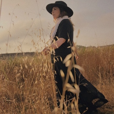 Mulher com vestido longo preto e chapéu preto em campo com ervas secas