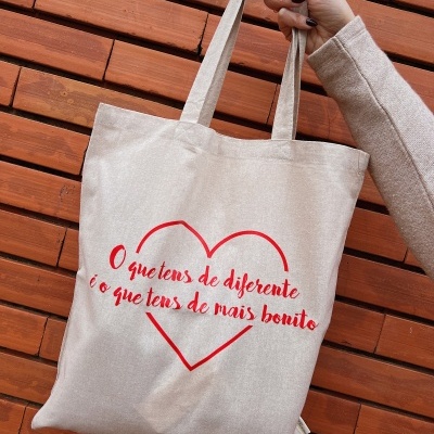 Saco de pano bege com texto vermelho e coração, segurado por mão com fundo de tijolos.