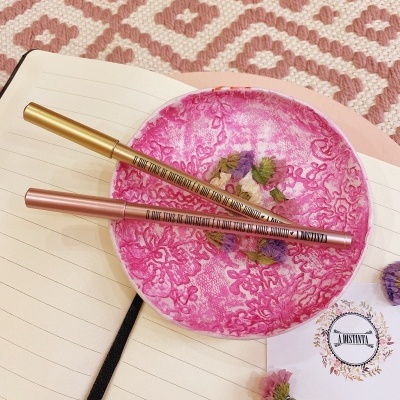 Canetas metálicas sobre minicumbuca rosa com flores secas, caderno aberto e cartão 'A DUSTINTA'