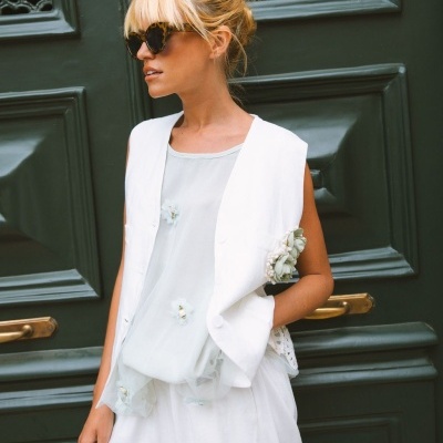 Mulher com colete branco e blusa transparente com flores, óculos de sol, ao lado de porta verde escura