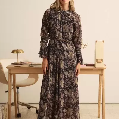 Mulher a usar vestido comprido floral e sapatos vinhos num escritório com secretária de madeira e cadeira bege.