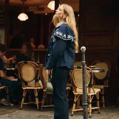Mulher com casaco azul bordado, calças escuras e bolsa verde junto a café com cadeiras de palhinha.